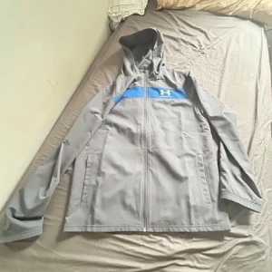 Grå vindjacka från Under Armour - Snygg grå vindjacka från Under Armour med blå detalj och logga på bröstet. Jackan har huva, hel dragkedja och två sidofickor. Tillverkad i polyester och perfekt för blåsiga dagar. Sportig look och bekväm passform.