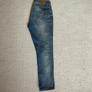 Blå jeans från Nudie Jeans, straight fit - Säljer ett par klassiska blå jeans från Nudie Jeans med snyggt slitna detaljer och kontrastsömmar. Modellen har raka ben och normal passform, med Nudie-logga på bakfickan och patch i midjan. Perfekta för dig som gillar en tidlös jeanslook. Längd 103-104cm