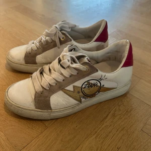 Zadig & Voltaire sneakers med blixt - Snygga sneakers från Zadig & Voltaire i vitt skinn med beige mockadetaljer, guldglittrig blixt på sidan och röd häl. Coolt grafittiliknande tryck vid sidan och klassisk snörning. Använda där av priset. Storlek 37. Pris är diskuterbart. 