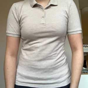 Beige pikétröja från Uniqlo, S - Snygg beige pikétröja från Uniqlo med klassisk krage och knappar framtill. T-shirten är i mjuk bomullsblandning med korta ärmar och figurnära passform. Perfekt för en clean och stilren look.