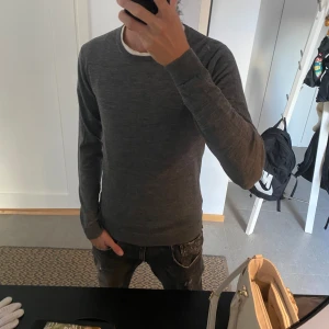 Merinoull tröja - Mörkgrå stickad tröja från Selected Homme i storlek S. Tröjan har rund halsringning, långa ärmar och en slim passform. Perfekt för lager-på-lager och funkar året runt. Enkel och stilren design som passar till det mesta. Två jättesmå hål som inte märks en på ryggen och en vid handleden.