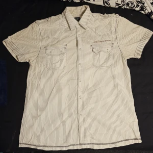 Beige randig skjorta med kort ärm - Kortärmad skjorta från MQ i beige med tunna svarta ränder. Skjortan har två bröstfickor med lock och knappar, klassisk krage och axelklaffar. Perfekt för en avslappnad men ändå stilren look.