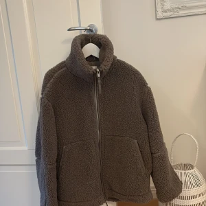  Brun teddyjacka från H&M, stl S - Mysig brun (mullvad) teddyjacka från H&M i storlek S. Jackan har hög krage, två stora fickor framtill och stängs med dragkedja. Tillverkad i 100% polyester med fluffig yta som ger en cool och avslappnad vibe. Perfekt för kyliga dagar. Köpt nyligen här på Plick, men stor i storlek så vi ska köpa XS istället! Denna är i jättefint skick och aldrig använd av oss!