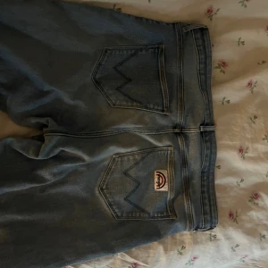 Wrangler ljusblå bootcut jeans - Säljer ett par ljusblå jeans från Wrangler med klassiska bakfickor och logga. Jeansen har bootcut-modell med utsvängda ben och hög midja. Tillverkade i jeansmaterial med snygg tvätt och raka sömmar. Perfekta för dig som gillar retro vibe och bekväm passform.