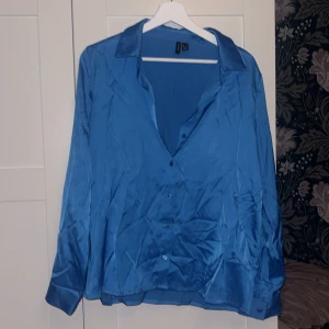 Blå skjorta från Vero Moda, storlek L - Snygg blå skjorta från Vero Moda i storlek L. Skjortan har klassisk krage, knappar framtill och långa ärmar. Materialet har en lätt glansig finish som ger en fräsch look. 