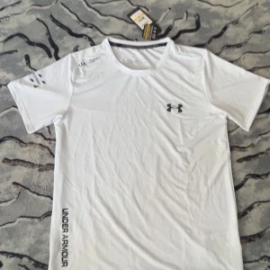Vit Under Armour t-shirt HeatGear - Snygg vit t-shirt från Under Armour med svart logga på bröstet och tryckt text längs sidan. Tillverkad i lätt och ventilerande polyester, perfekt för träning. Klassisk rund hals och korta ärmar. Modellen är slim och passar dig som gillar sportig stil.