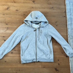 Grå hoodie från Sail Racing med dragkedja - Grå hoodie från Sail Racing med hel dragkedja framtill och huva. Insidan är fodrad med mjuk fleece för extra komfort. Tröjan har två fickor med dragkedja och Sail Racing-logga på bröstet. Perfekt för chill dagar och sköna promenader.
