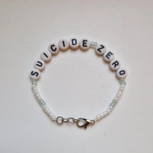 Egengjort pärlarmband Suicide Zero - Handgjort armband med vita och ljusgröna pärlor samt bokstavspärlor som bildar texten 'SUICIDE ZERO'. Armbandet har ett smidigt metallspänne och är ca 15-17 cm långt men är också gjort med töjbar tråd. Perfekt accessoar för att visa stöd och sprida budskap. 