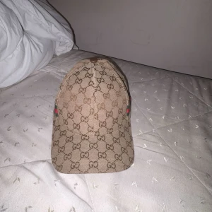 Gucci keps med GG-mönster och röd/grön detalj - Snygg beige keps från Gucci med klassiskt GG-monogram över hela ytan. Kepsen har en böjd skärm och en ikonisk röd och grön rand på sidan. Tillverkad i bomullsmix med justerbar rem baktill. Perfekt för dig som vill sticka ut med en lyxig accessoar.