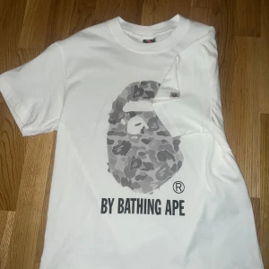 BAPE By Bathing Ape T-shirt Vit  - Cool vit T-shirt från BAPE (A Bathing Ape) med klassiskt camo-apehead tryck på bröstet och “By Bathing Ape” text under. ✔️ Storlek: M/S ✔️ Färg: Vit med grått camotryck
