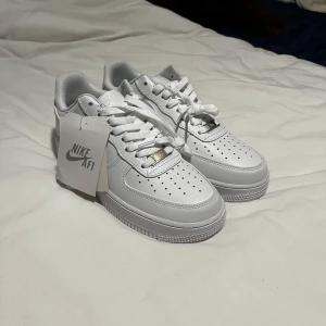 Nike Air Force 1 vita sneakers - Klassiska Nike Air Force 1 sneakers i helvitt skinn med perforerad tå, rund tå och platt sula. Snygg och tidlös modell med snörning och diskreta silverdetaljer. Perfekta för dig som gillar en clean och stilren look.