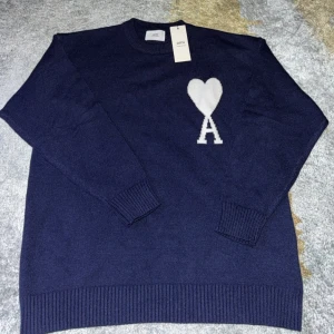 Mörkblå stickad tröja från Ami - Mörkblå stickad tröja från Ami Alexandre Mattiussi med vit hjärtlogga och bokstaven A på bröstet. Klassisk rund halsringning och ribbade muddar vid ärmslut och nederkant. Perfekt för dig som gillar stilrena och trendiga plagg.