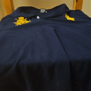 Mörkblå pikétröja från Polo Ralph Lauren - Klassisk mörkblå pikétröja från Polo Ralph Lauren med gul broderad logga på bröstet. Tröjan har krage, knäppning med två knappar och är tillverkad i mjuk bomull. Perfekt för en stilren och sportig look.