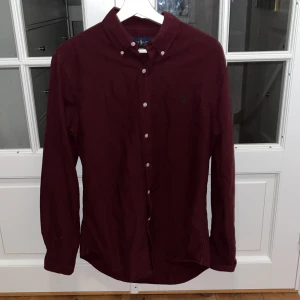 Vinröd skjorta från Ralph Lauren - Vinröd skjorta från Ralph Lauren i slim fit med klassisk button-down krage och broderad logga på bröstet. Skjortan har långa ärmar och vita knappar framtill. Tillverkad i bomull, perfekt för dig som gillar stilrena och snygga plagg.