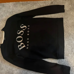 Svart sweatshirt från Hugo Boss - Svart sweatshirt från Hugo Boss med stor silverfärgad logga framtill. Klassisk rund halsringning och långa ärmar. Perfekt för dig som gillar stilrena och trendiga plagg med tydlig branding.