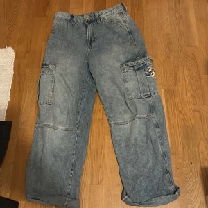 Baggy cargojeans med fickor - Säljer ett par baggy cargopants i ljusblå denim med stora sidofickor och cool patch på ena benet. Byxorna har bred passform och är perfekta för en avslappnad streetstil. Jeansen har synliga sömmar och klassisk cargodesign.