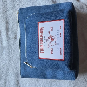 True Religion jeansväska med patch - Snygg jeansväska från True Religion med stor vit och röd patch framtill. Väskan har två fack med dragkedja och är tillverkad i blått jeanstyg med kontrastsömmar. Perfekt för dig som gillar streetstyle och vill sticka ut med en unik accessoar.