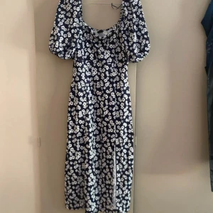 Blommig långklänning från Gina Tricot - Supersöt långklänning från Gina Tricot med marinblå botten och vita blommor. Klänningen har puffiga kortärmar, rund halsringning och en snygg slits framtill. Perfekt för sommarens alla tillfällen!