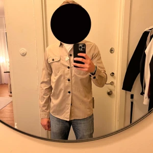 Beige overshirt från Matinique (Nypris 1299) - Stilren beige overshirt från Matinique med svarta knappar och två bröstfickor. Skjortan är i bomull och har en klassisk, rak passform med lång ärm. Perfekt att bära öppen över en t-shirt för en avslappnad look. Helt ny, aldrig använd. Nypris 1299