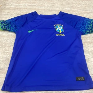 Blå Brasilien fotbollströja Nike - Snygg blå Brasilien fotbollströja från Nike med grönt märke och CBF-logga på bröstet. Ärmarna har leopardmönster i blått och grönt. Tillverkad i lätt Dri-FIT-material som andas. Perfekt för dig som älskar fotboll och vill sticka ut på planen.