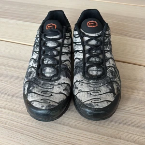 Nike Air Max Plus svart/vit EUR 39 - Nike Air Max Plus sneakers i svart och vit med unikt mönster och svarta vågformade detaljer. Skorna har svart sula, snörning och orange Nike-logga på sidan och plös. Ovandelen är i textil och syntet, perfekt för dig som gillar streetstyle. Ny pris 2800kr säljs för 800kr. 