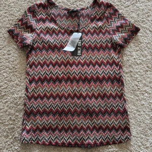 Mönstrad omlottopp från ONLY  - Färgstark omlottopp från ONLY med kort ärm och coolt sicksackmönster i rosa, rött, svart, beige och guld. Toppen har omlottdetalj baktill och är tillverkad i ett glittrigt, stretchigt material som ger en unik look.