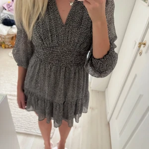 Svartvit kortklänning med volang - Supersöt kortklänning i svartvitt småmönstrat tyg med v-ringning, smockad midja och volangkant nertill. Klänningen har trekvartsärmar i luftig, lätt transparent polyester och en flowy passform som känns både trendig och bekväm.