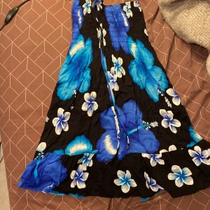 Blommig blå och svart sommarklänning - Säljer en kort, axelbandslös klänning med stora blå och vita blommor på svart botten. Klänningen har smock upptill och volang nertill, perfekt för varma dagar. Lätt och luftig modell med knytband framtill.