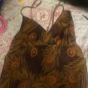 Brun linne med påfågelfjädrar Gina - Supersnygg brun linne från Gina med tunna axelband och v-ringning. Linné har ett unikt mönster av påfågelfjädrar i gult och orange, vilket ger en cool och bohemisk vibe. Perfekt för dig som vill sticka ut med ett färgstarkt plagg.