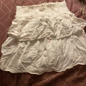 Vit blommig volangkjol med resår - Supersöt vit kortkjol med små blommor i rosa och blått. Kjolen har bred smockresår i midjan och dubbla volanger för extra flowy känsla. Perfekt för dig som gillar romantisk och feminin stil.