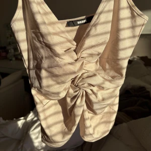 Beige ribbad linnetopp från BikBok - Supersnygg beige linnetopp från BikBok med ribbad struktur och twistad knut framtill. Toppen har smala axelband och v-ringning, vilket ger en trendig och fräsch look. Perfekt att styla med jeans eller kjol för en cool vibe.