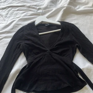 Svart omlottopp från Gina Tricot - Svart omlottopp från Gina Tricot med lång ärm och v-ringning. Toppen har knytband i midjan och är tillverkad i ett lätt, strukturerat tyg som ger en snygg passform. Perfekt att styla med jeans eller kjol för en trendig look.