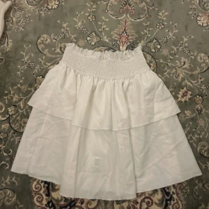 Vit volangkjol med smock i midjan - Supersöt vit kortkjol med volanger och smockad midja. Kjolen har en luftig känsla och är perfekt för varma dagar. Den är tillverkad i bomull och har en feminin, flowy look som är lätt att matcha med olika toppar. ingen aning vart jag köpte den