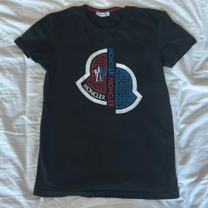 Svart Moncler t-shirt med tryck - Svart t-shirt från Moncler i storlek S med stort grafiskt tryck i rött, blått och vitt på bröstet. Klassisk rund hals och korta ärmar. Den är gjord av 100% bomull.