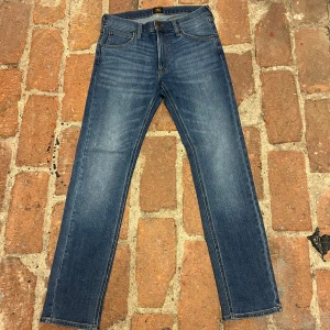 Lee Jeans - Klassiska blå jeans från Lee med raka ben och femficksmodell. Jeansen har mellanblå tvätt, subtila slitningar och Lee-läderpatch bak i midjan. Tillverkade i slitstarkt denimtyg med snygga kontrastsömmar. Perfekta för en avslappnad och stilren look.