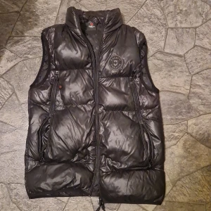 Svart dunväst från Zavetti Canada - Svart dunväst från Zavetti Canada med hög krage och två fickor med dragkedja. Västen har ett stilrent emblem på bröstet och blanka ytor som ger en modern look. Perfekt för lager-på-lager och har en normal passform. 