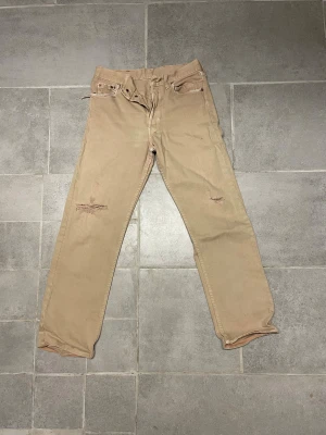 Beige Levi's 501 jeans W28 L30 - Säljer ett par klassiska Levi's 501 jeans i beige med raka ben och fem fickor. Jeansen har slitna detaljer och är tillverkade i bomull. Passar dig som gillar en tidlös och avslappnad stil. Made in UK.