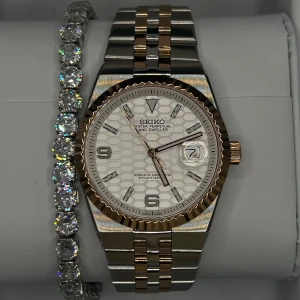 Seiko Land-Dweller tvåtonad automatisk klocka - Säljer en Seiko Land-Dweller automatisk klocka med tvåtonad länk i silver och guld. Urtavlan är vit med hexagonmönster, datumfönster och lysande visare. Boetten har räfflad bezel i guldton och transparent baksida som visar urverket. Perfekt för dig som gillar stil och teknik.