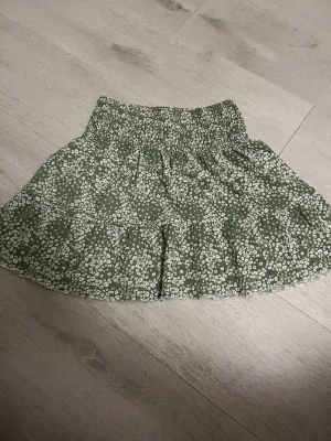 Grön blommig volangkjol Gina Tricot - Supersöt grön kortkjol från Gina Tricot med vitt blommönster och volangdetaljer. Kjolen har smockad midja som ger en bekväm och snygg passform. Perfekt för dig som gillar en feminin och trendig stil. Passar XS/S