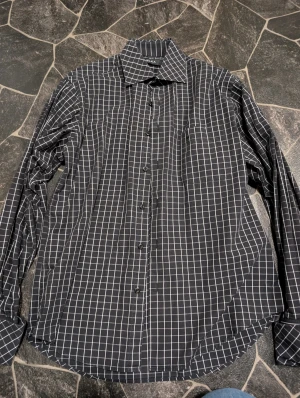 Svart rutig skjorta från Melka stl M  - Svart skjorta från Melka i storlek M med vitt rutmönster och klassisk krage. Skjortan har långa ärmar, knäppning framtill och en bröstficka. Perfekt för dig som gillar stilren och enkel design. Man behöver machett knappar till denna skjorta