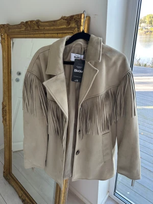 Beige mockajacka med fransar Rita Ora - Snygg beige mockajacka från Rita Ora x Primark med långa fransar över bröst, rygg och ärmar. Jackan har klassisk krage, knäppning framtill och två fickor. 