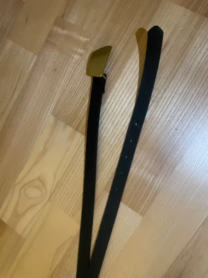 Svart skärp med guldfärgat spänne - Stilrent svart bälte med ett snyggt guldfärgat spänne. Bältet är smalt och har en klassisk, rak form som passar till jeans eller kostymbyxor. Materialet är skinn och designen är enkel utan mönster eller logga. Perfekt för dig som gillar minimalistisk stil.