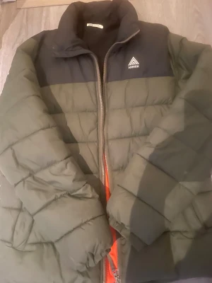 Grön pufferjacka från NWRK - Snygg pufferjacka från NWRK i olivgrönt med svarta axelpartier och orange innerfoder. Jackan har hög krage, quiltad design och dragkedja framtill. Perfekt för kalla dagar och ger en cool streetstyle-vibe.