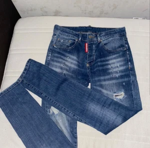 Dsquared2 blå jeans med slitningar - Säljer ett par snygga Dsquared2 jeans i blå tvätt med coola slitningar och distressed detaljer. Jeansen har rakt snitt, klassisk femficksdesign och Dsquared2-logga på röd etikett fram samt unik patch bak. Perfekt för dig som gillar streetwear och vill sticka ut.