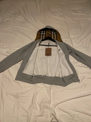 Grå Burberry hoodie - Helt ny grå hoodie från Burberry med klassisk rutig huva i beige, svart och röd. Tröjan har dragkedja framtill, snörning vid huvan och broderad Burberry-logga på bröstet. Tillverkad i mjuk bomull för skön komfort och har långärmad passform.