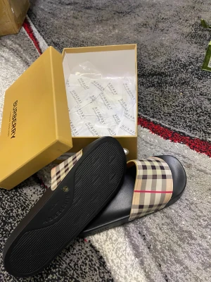 Burberry sandaler med klassiskt rutmönster - Snygga sandaler från Burberry med den ikoniska rutan i beige, svart, röd och vit på remmen. Sulan är svart och har Burberry-logga undertill. Perfekt för sommaren och riktigt stilrena att glida runt i. Kommer med originalkartong och påse.