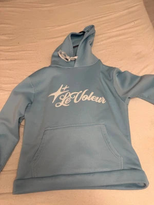 Ljusblå Le Voleur hoodie med tryck - Säljer en ljusblå hoodie från Le Voleur i storlek M. Tröjan har ett stort vitt tryck på bröstet, snörning i huvan och en klassisk magficka. Materialet är mjukt och fleece-liknande, perfekt för chill dagar. Snygg och enkel design med lång ärm och relaxed passform.