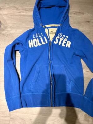 Blå hoodie från Hollister med dragkedja - Snygg blå hoodie från Hollister med broderad vit logga och text på bröstet. Tröjan har huva med snörning, praktiska fickor framtill och dragkedja. Mjuk insida i fleece och ribbade muddar vid ärmslut och nederkant. Perfekt för chill eller häng.