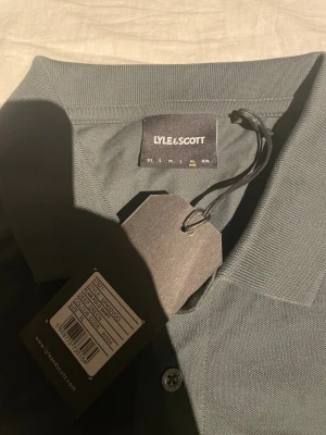 Grön pikétröja från Lyle & Scott XL - Snygg grön pikétröja från Lyle & Scott i storlek XL. Klassisk krage, korta ärmar och knappar framtill. Tröjan har det ikoniska gula Lyle & Scott-logotypen broderat på bröstet. Tillverkad i mjuk bomull som känns skön mot huden.
