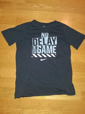 Svart Nike t-shirt med tryck - Svart t-shirt från Nike i storlek L med stort vitt tryck på bröstet där det står 'NO DELAY IN MY GAME'. Klassisk rund hals och korta ärmar. Tillverkad i 100% bomull för skön känsla och bra passform. Perfekt för dig som gillar sportig stil.